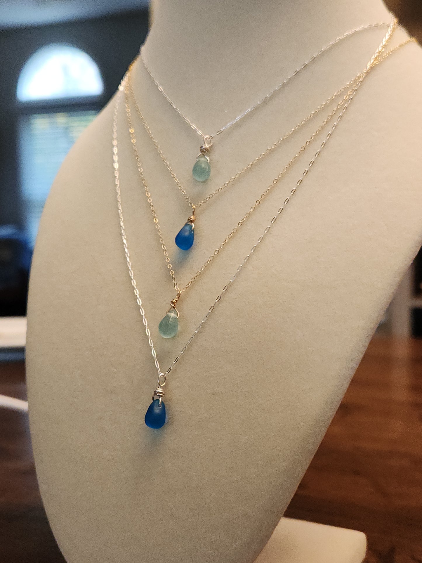 Layered gold-filled necklaces displayed on a jewelry bust, featuring light and dark blue sea glass pendants in varying lengths.