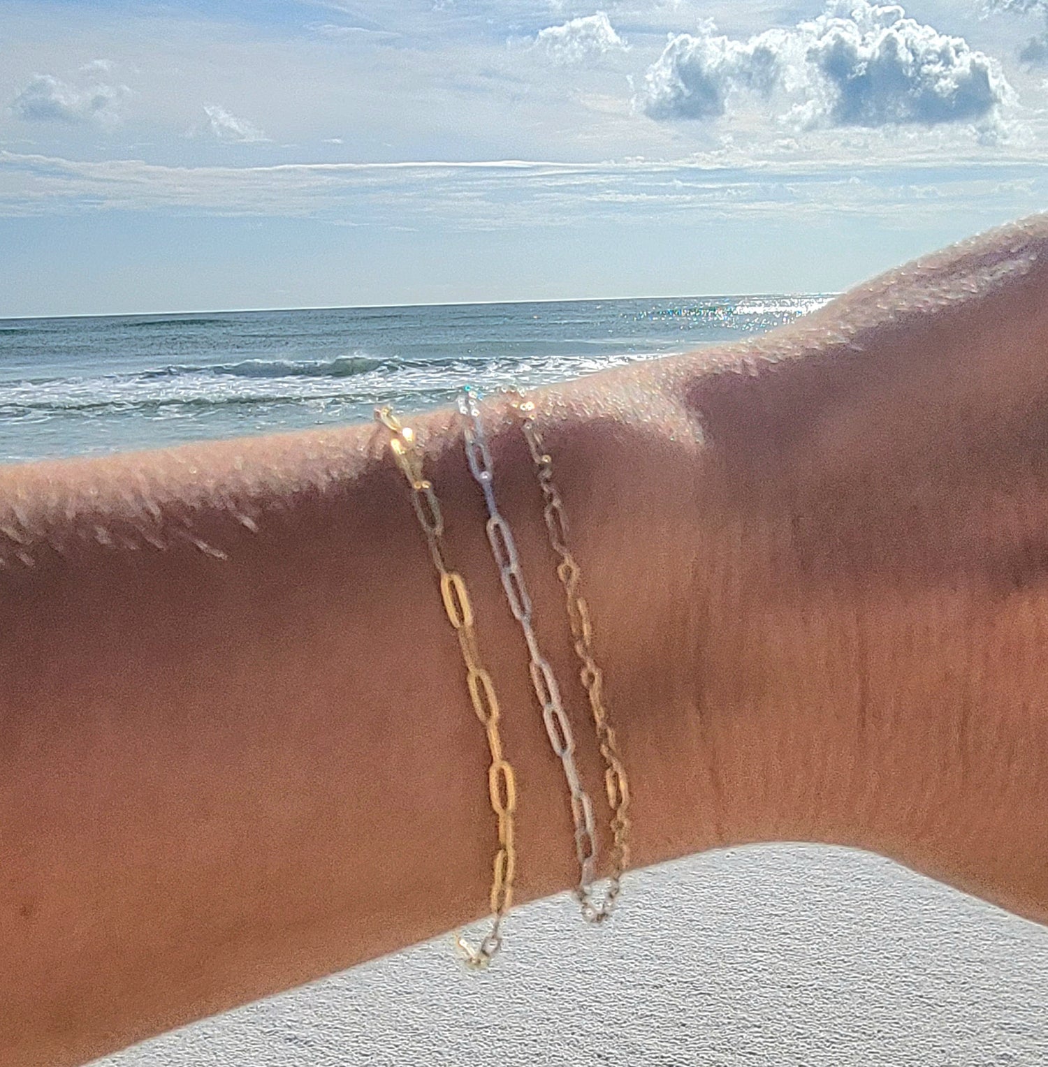 Permanent jewelry bracelets in 14k gold-filled and sterling silver chains worn on a wrist with an ocean beach background