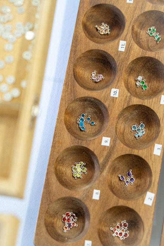 Round birthstone charms for permanent jewelry displayed in a wooden tray, available in multiple gemstone colors with updated photos coming soon
